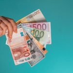 A hand holding a fan of assorted Euro and US dollar banknotes against teal