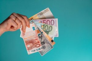 A hand holding a fan of assorted Euro and US dollar banknotes against teal