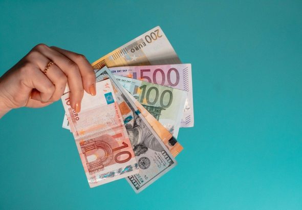 A hand holding a fan of assorted Euro and US dollar banknotes against teal